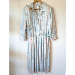 May include: A midi-length dress featuring a light blue, pink, and white swirling paisley pattern. The dress has a collared neckline, a single gold-toned button closure, three-quarter length sleeves with cuffs, and an elasticized waistline.