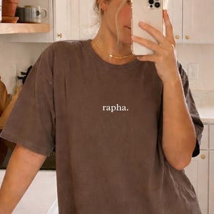 Può includere: Una maglietta marrone con la scritta "rapha" stampata sul davanti. La maglietta è indossata in un ambiente cucina con mobili bianchi e mensole in legno. La persona tiene un telefono davanti al viso.