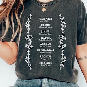 Puede incluir: Una camiseta gris oscuro con un diseño blanco que presenta los nombres de Dios en hebreo con sus significados en inglés. El diseño está enmarcado por dos vides blancas con flores.