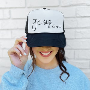 K&ouml;nnte beinhalten: Eine schwarz-wei&szlig;e Trucker-Kappe mit dem Text "Jesus is King" in schwarzer Schrift auf der Vorderseite.