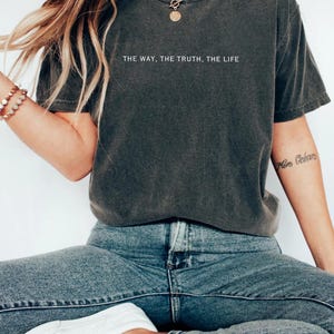Może przedstawiać: Szary t-shirt z nadrukiem "THE WAY, THE TRUTH, THE LIFE" na przodzie. Koszulkę nosi osoba w niebieskich dżinsach.
