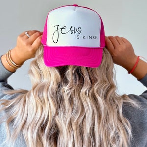 K&ouml;nnte beinhalten: Eine rosafarbene und wei&szlig;e Trucker-Kappe mit dem Text "Jesus is King" in schwarzer Schrift.