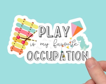 Puede incluir: Una pegatina blanca con el texto "PLAY is my favorite OCCUPATION" en negro. La pegatina presenta ilustraciones coloridas de un xilófono, una cometa y un dinosaurio.