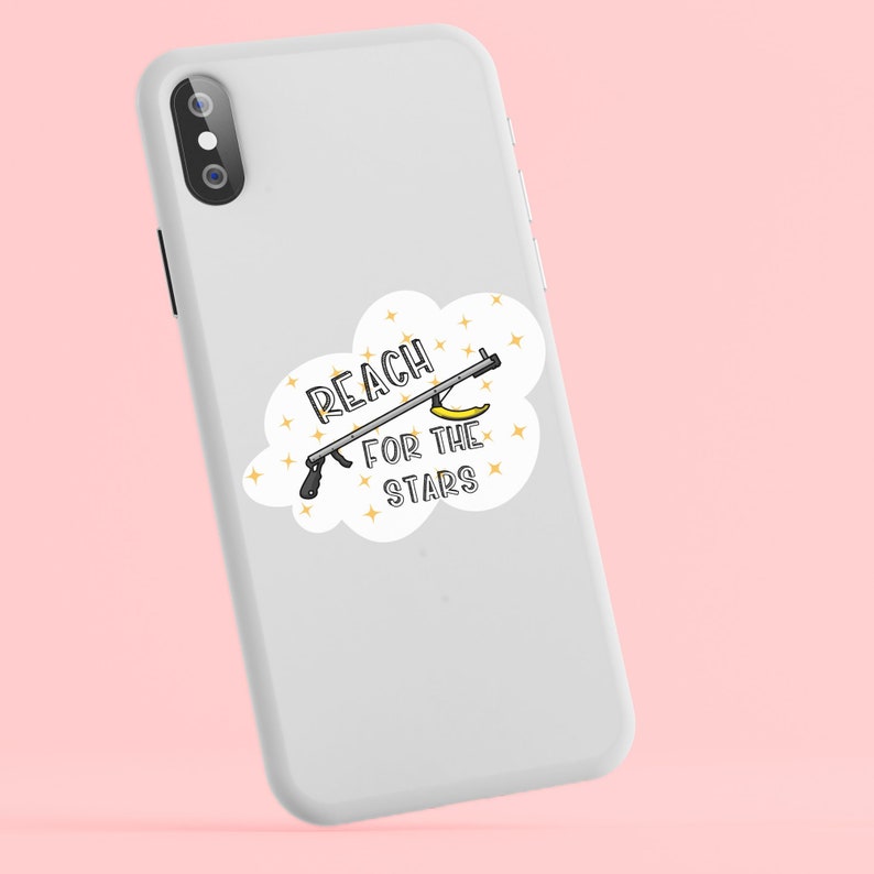 Puede incluir: Una funda de tel&eacute;fono blanca con una pegatina que dice "Reach for the stars" y muestra un pl&aacute;tano sostenido por una herramienta con un mango en forma de estrella. La pegatina est&aacute; rodeada de estrellas doradas.