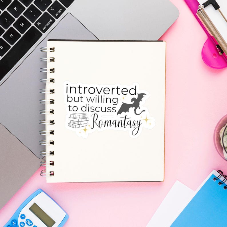May include: A white notebook with a sticker that says "introverted but willing to discuss Romantasy" with a black silhouette of a dragon and a stack of books.