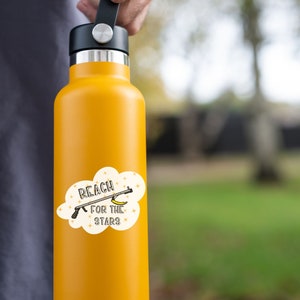 Puede incluir: Una botella de agua amarilla con una pegatina blanca que dice "Reach for the Stars" y presenta una pistola de pl&aacute;tano de dibujos animados.
