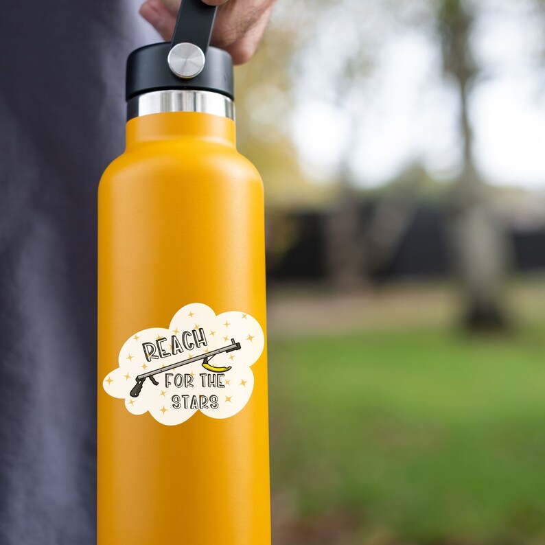 Puede incluir: Una botella de agua amarilla con una pegatina blanca que dice "Reach for the Stars" y presenta una pistola de pl&aacute;tano de dibujos animados.