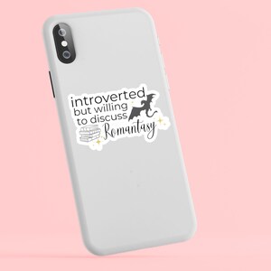 May include: White iPhone case with a black and white sticker that says "introverted but willing to discuss romantasy" with a silhouette of a dragon and a stack of books.