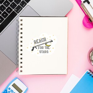 Puede incluir: Un cuaderno blanco con una encuadernaci&oacute;n en espiral y una pegatina que dice "Reach for the stars" con una imagen de dibujos animados de un pl&aacute;tano en un palo. La pegatina est&aacute; sobre un fondo rosa.