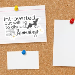 May include: A white sticker with black text that reads "introverted but willing to discuss romantasy" with a black silhouette of a dragon and a stack of books. The sticker is on a corkboard with two white pieces of paper.