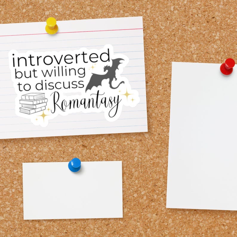 May include: A white sticker with black text that reads "introverted but willing to discuss romantasy" with a black silhouette of a dragon and a stack of books. The sticker is on a corkboard with two white pieces of paper.