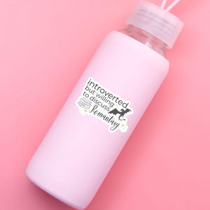 May include: A pink water bottle with a white sticker that says "introverted but willing to discuss Romantasy". The sticker features a stack of books, a dragon, and stars.