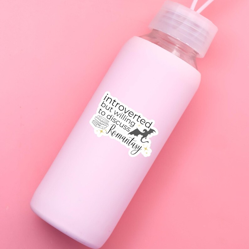 May include: A pink water bottle with a white sticker that says "introverted but willing to discuss Romantasy". The sticker features a stack of books, a dragon, and stars.