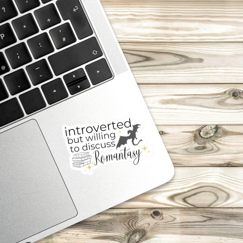 May include: A white sticker with black text that reads "introverted but willing to discuss Romantasy" with a black silhouette of a dragon and a stack of books.
