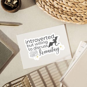 May include: A white card with the text "introverted but willing to discuss Romantasy" in black lettering. A black silhouette of a dragon is flying above a stack of books. The card is on a white surface.