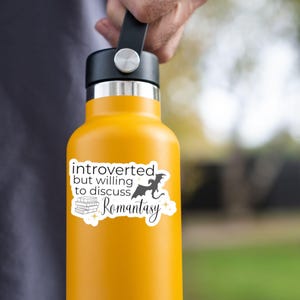 May include: A yellow water bottle with a white sticker that says "introverted but willing to discuss Romantasy" with a black dragon and a stack of books.
