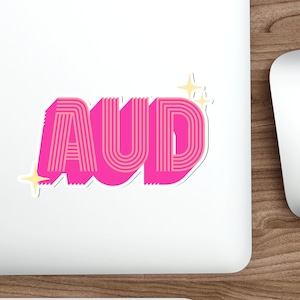 May include: A sticker with the letters "AUD" in a retro font. The letters are bright pink with a striped design and a white outline. Two yellow starbursts are on the left side of the sticker.