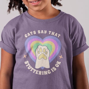 May include: A young person wearing a purple t-shirt with a rainbow heart and paw print graphic. The text on the shirt reads "Cats Say That Stuttering Is Ok."