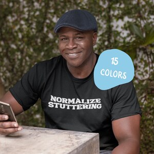 May include: A person wearing a black t-shirt with the text "Normalize Stuttering" in white letters. The person is wearing a dark blue cap and is holding a black smartphone. The background is a blurred image of green foliage. A blue speech bubble with the text "15 Colors" is in the upper right corner of the image.