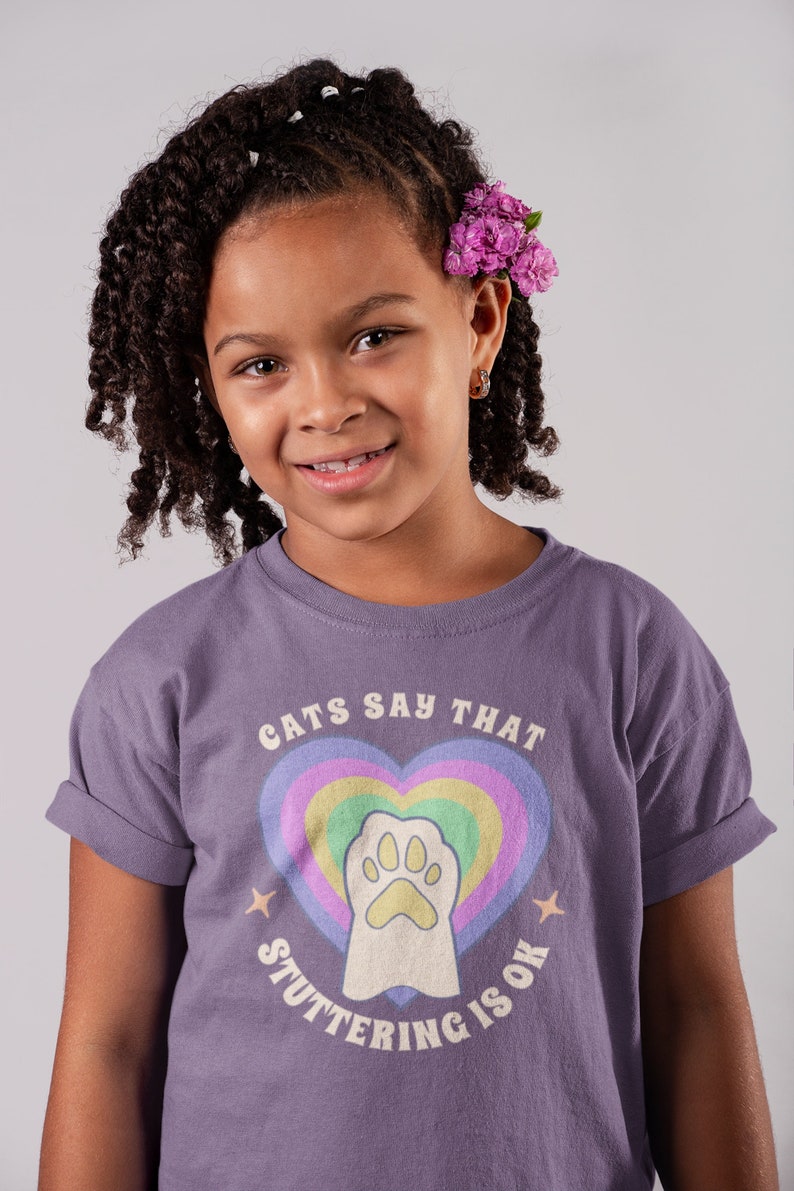 May include: A young person wearing a purple t-shirt with a rainbow heart and paw print graphic. The text on the shirt reads "Cats Say That Stuttering Is Ok."