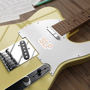 Pu&ograve; includere: Una chitarra elettrica gialla con un adesivo floreale che dice "SLIP" sul corpo. La chitarra ha un battipenna cromato e un pickup nero.