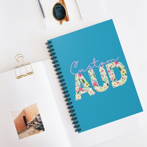 May include: A blue spiral notebook with the text "Custom AUD" in pink and white lettering. The letters are filled with a floral pattern.