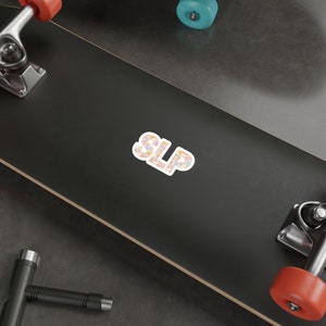 Pu&ograve; includere: Uno skateboard nero con un adesivo bianco che dice "SLP" in lettere floreali rosa, arancio e gialle. L'adesivo &egrave; sulla parte superiore dello skateboard.