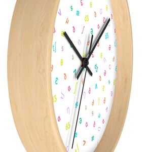 May include: A white clock face with black hands and a wooden frame. The clock face is covered in a random pattern of colorful letters and symbols.