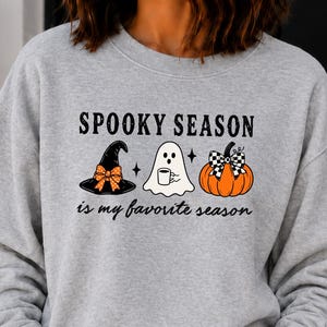 Pode incluir: Uma camisola cinzenta clara de gola redonda com o texto preto SPOOKY SEASON is my favorite season. O gráfico inclui um chapéu de bruxa preto com um laço laranja às bolinhas, um fantasma branco a segurar uma caneca e uma abóbora laranja com um laço axadrezado preto e branco.