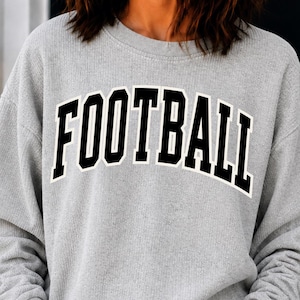 Puede incluir: Una sudadera de cuello redondo y manga larga en color gris claro con la palabra FOOTBALL impresa en una fuente negra, arqueada y negrita de estilo universitario con un borde blanco en el pecho. La prenda tiene un acabado de tela acanalada.