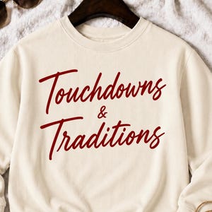 Puede incluir: Una sudadera de cuello redondo color crema con el texto granate Touchdowns & Traditions impreso en la parte delantera. La prenda tiene mangas largas, cuello acanalado y puños acanalados. El diseño está centrado en la zona del pecho.