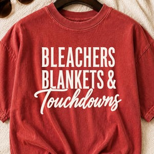 May include: A red short-sleeved t-shirt with a crew neckline featuring the white printed text BLEACHERS BLANKETS & Touchdowns. The garment has a washed, vintage-style appearance and is displayed on a black hanger against a textured white fabric.