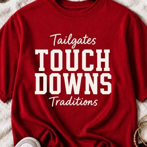 Może przedstawiać: Czerwony t-shirt z krótkim rękawem z białym napisem Tailgates, TOUCH DOWNS oraz Traditions. Koszulka ma okrągły dekolt, jest zawieszona na czarnym wieszaku i posiada wyrazisty nadruk graficzny umieszczony na środku z przodu.