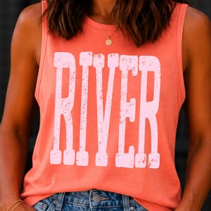 Könnte beinhalten: Korallenfarbenes Tanktop mit dem Wort "RIVER" in großen, weißen Buchstaben im Used-Look. Das Top ist ärmellos und wird mit einer blauen Jeans kombiniert. Eine goldene Halskette und ein Armband sind sichtbar.
