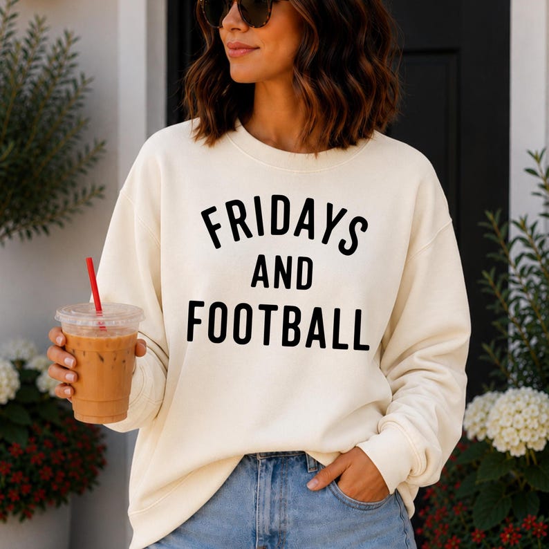 May include: A cream-colored crewneck sweatshirt featuring the black text FRIDAYS AND FOOTBALL printed in a bold, sans-serif font across the chest. The garment has long sleeves with ribbed cuffs and a ribbed neckline, paired with blue denim jeans.