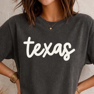 May include: Dark gray t-shirt with the word "Texas" in a white, cursive font. The shirt has a crew neck and short sleeves. The image is a close-up of the shirt, showing the text prominently.