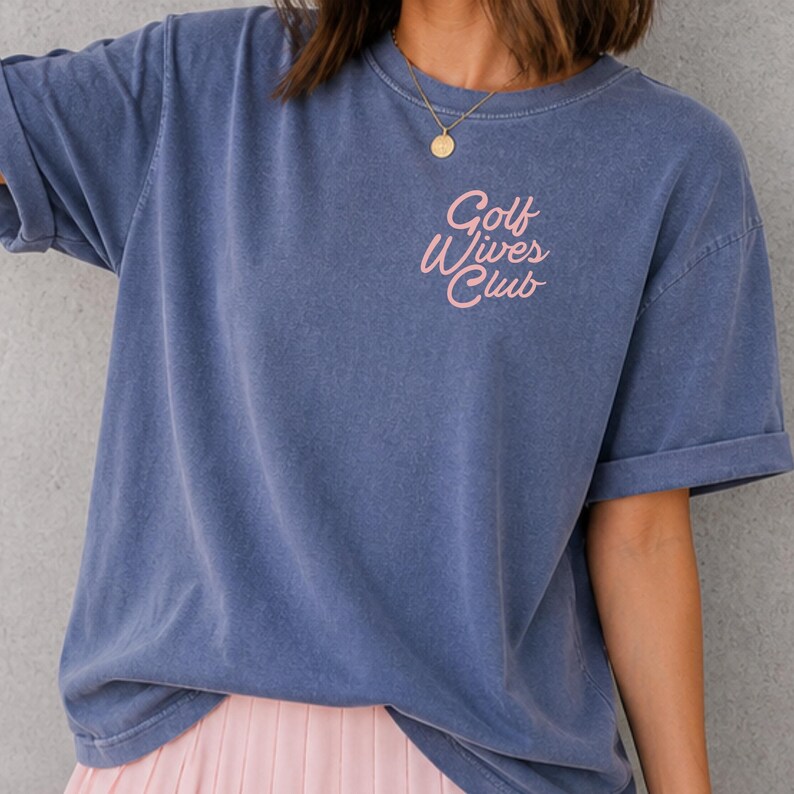 May include: A blue short-sleeved t-shirt with the text "Golf Wives Club" in pink script. The shirt has a relaxed fit and rolled-up sleeves. The model is wearing a gold necklace.