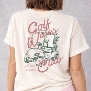 May include: Cream-colored t-shirt with a pink and green graphic of a golf cart and the words "Golf Wives Club." The text "good times & tee times" is below the cart. The shirt has short sleeves and a relaxed fit.