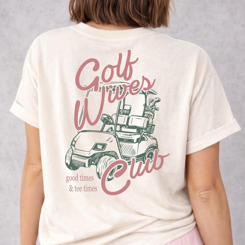 May include: Cream-colored t-shirt with a pink and green graphic of a golf cart and the words "Golf Wives Club." The text "good times & tee times" is below the cart. The shirt has short sleeves and a relaxed fit.