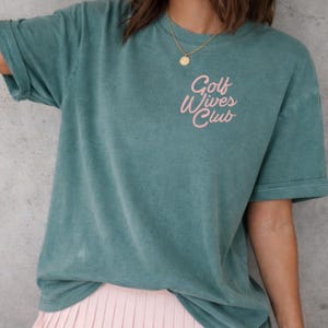May include: A teal-colored t-shirt with the text "Golf Wives Club" in pink script. The shirt has short sleeves and a relaxed fit. A gold necklace is visible at the neckline. The shirt is paired with a pink skirt.