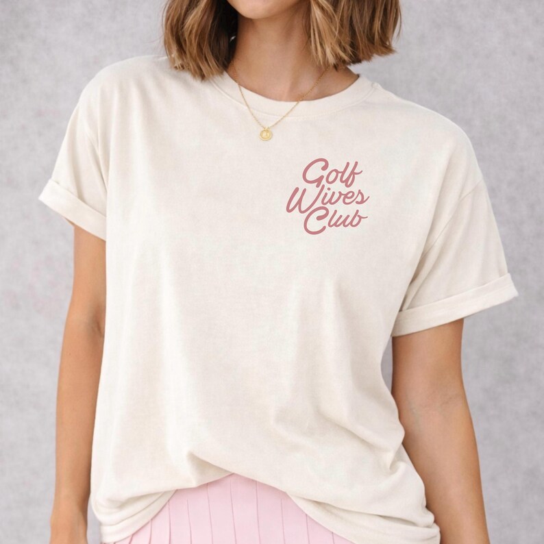 May include: Cream-colored t-shirt with rolled-up sleeves. The shirt features the text "Golf Wives Club" in a cursive, reddish-pink font on the upper left chest. A gold necklace is visible.