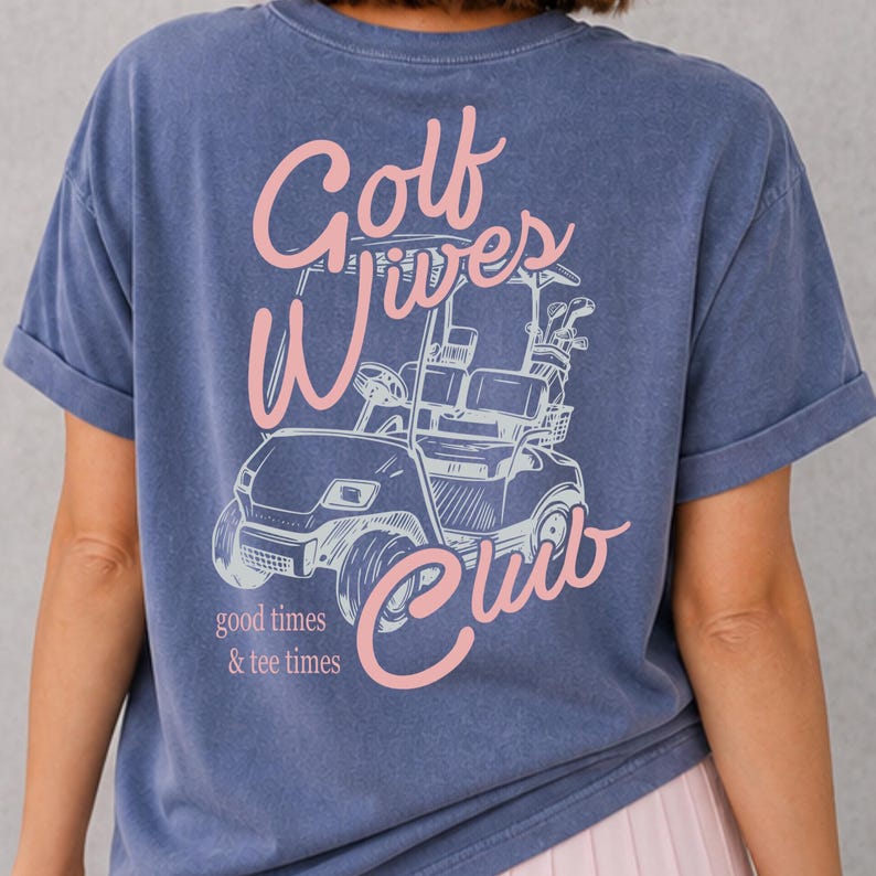 May include: Blue t-shirt with a pink graphic of a golf cart and the words "Golf Wives Club." The text "good times & tee times" is also printed on the shirt. The shirt has rolled-up sleeves.