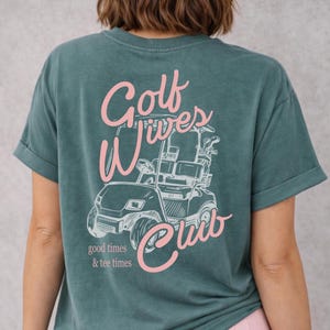May include: Teal-colored t-shirt with a graphic of a golf cart and the words "Golf Wives Club" in pink script. The phrase "good times & tee times" is printed below the cart.
