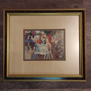 May include: A framed print of a colorful painting depicting people sitting at a table outdoors. The painting features a vibrant palette of blues, greens, yellows, and reds. The people are dressed in various styles, and the scene is set in a garden or park setting.
