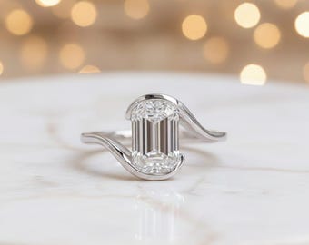 May include: A silver ring with a large, rectangular, emerald-cut diamond. The band features a unique, flowing design that curves around the diamond. The ring is set on a white marble surface, with a blurred background of warm, golden lights.