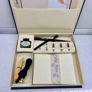 Op de afbeelding: Een open doos met een set voor kalligrafie met een zwarte inktfles, vijf penpunten, een zwarte penhouder, een zegel van was, een zegel van was en een stapel blanco briefpapier met een bloemenmotief.