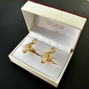 Puede incluir: Dos anillos de clave de sol de oro en una caja blanca con interior rojo. La caja tiene el texto "EPNKY 2. @EZ/NIKH THAI 2910 541.908" impreso en el interior.