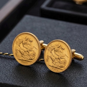 Puede incluir: Un par de gemelos de color dorado con un diseño de moneda circular. Cada moneda muestra un relieve de una figura a caballo matando a un dragón, con el año 1916 inscrito en la parte inferior. Los gemelos tienen un acabado metálico pulido.
