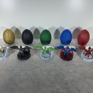 May include: Seven dragon figurines, each with a different colored dragon egg, on a white surface. The dragons are all in a flying pose with their wings spread out. The eggs are gold, black, green, blue, and red.