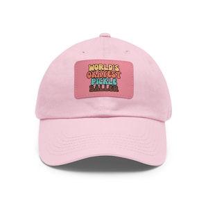 May include: Pink baseball cap with a patch that says "WORLD'S OKAYEST PICKLE BALLER" in a retro font with colourful letters.